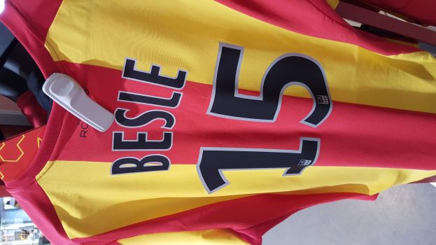 Rotes und gelbes Fußballtrikot mit Text und Zahlen, das auf einem Ständer in einem Geschäft hängt, zusammen mit anderen Gegenständen und dem Boden im Hintergrund.