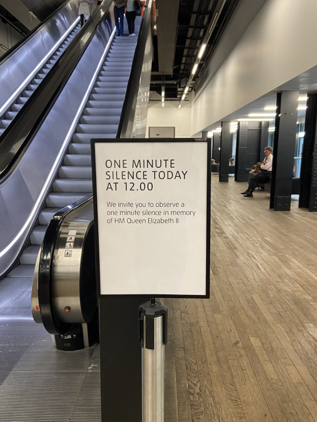 Eine Rolltreppe in einem Flughafen mit einem Schild, auf dem "Eine Minute Stille heute" steht, einige Menschen darauf und Lichter an der Decke im Hintergrund.