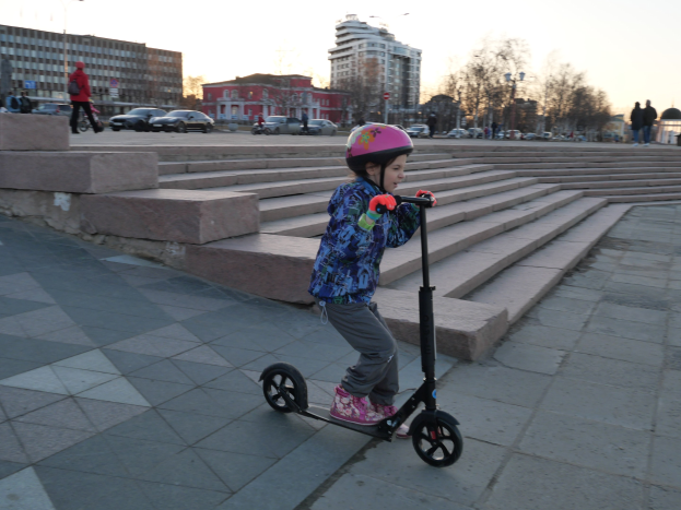 Ein junges Mödchen in einem Helm fährt auf einem Scooter eine Treppe hinunter mit Fahrzeugen, Menschen, Laternenmästen, Bäumen, Gebäuden und einem klaren blauen Himmel im Hintergrund.