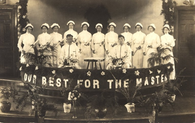 Eine Gruppe von Frauen in weißen Kleidern und Hauben steht auf einer Bühne mit einem Banner, auf dem 'Unser Bestes für den Meister' steht, umgeben von Blumentöpfen und Türen sowie einer Wand im Hintergrund.