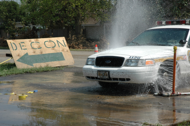 Ein Polizeiauto fährt durch eine Pfütze mit verstreuten Gegenständen, darunter ein Verkehrskegel und eine Tafel auf dem Boden, vor einem Hintergrund aus Bäumen, Gebäuden und einem klaren blauen Himmel.