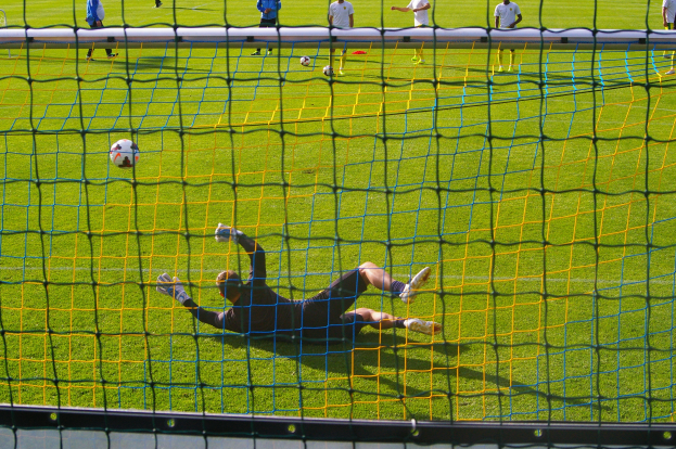 Ein Fussballspieler hält den Ball vor einem Tor, mit einem Netz im Vordergrund und einigen Menschen im Hintergrund sowie mehreren Bällen auf dem Boden verstreut.