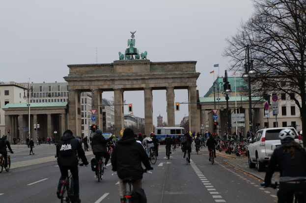 Eine Gruppe von Menschen, die auf Fahrrädern eine Straße vor dem Brandenburger Tor in Berlin, Deutschland, entlangfährt, mit Bäumen, Laternen, Verkehrszeichen, Schildern und Gebäuden mit Fenstern im Hintergrund und dem Himmel in der Ferne.