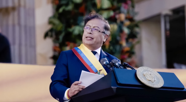 Ein Mann in einem blauen Anzug und Krawatte steht an einem Rednerpult, hält Papiere und spricht in ein Mikrofon, mit einer Pflanze und einer Wand im Hintergrund, was eine formelle Umgebung suggeriert.