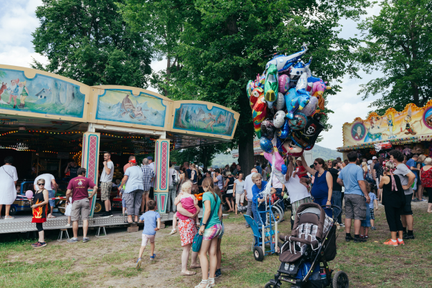 Eine Menschenmenge, einige mit Kinderwagen, umringt einen bunt beleuchteten Karussellwagen auf einem Volksfest, mit Bäumen und einem bewölktem Himmel im Hintergrund.