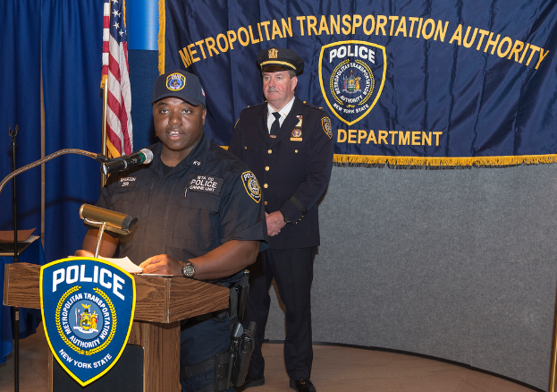Zwei Polizisten stehen vor einem Rednerpult, einer hält Papers und spricht in ein Mikrofon, mit einer Fahne, einem Tisch und einer Tafel mit Symbolen und Text im Hintergrund.