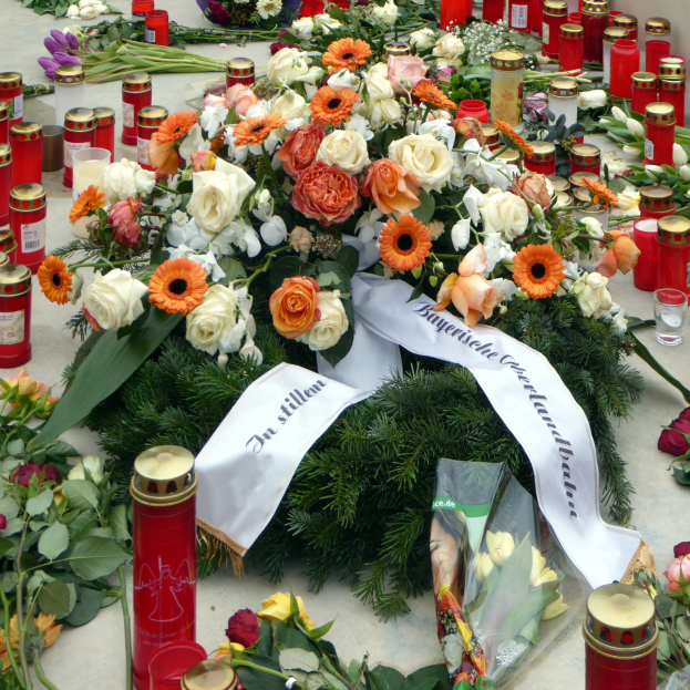 Ein Mahnmal in Berlin, Deutschland, mit einem Blumenstrauß und Kerzen, die auf dem Boden vor einer Wand aufgestellt sind, umgeben von Blättern und anderen Gegenständen.