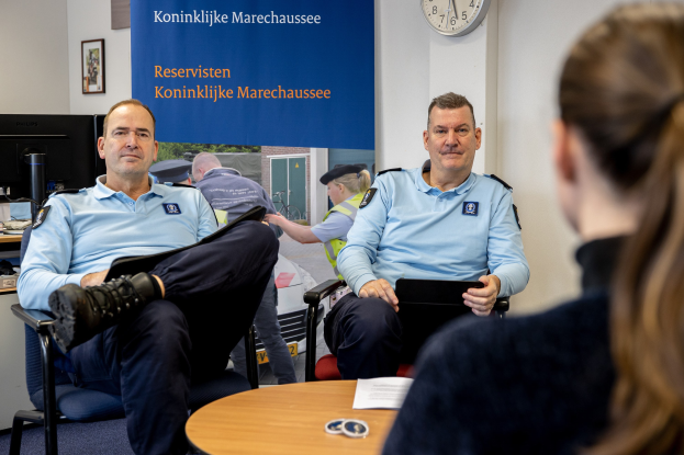 Zwei Polizeibeamte sitzen an einem Tisch mit Papieren und Gegenständen und sprechen mit einer Frau, mit einer Fahne, Uhr und Foto Rahmen an der Wand und einem weiteren Tisch mit Monitor und Tastatur links.