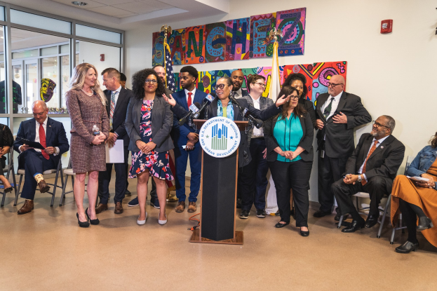 Gruppe von Menschen in einem Raum mit einem Podium, Mikrofon und einer Tafel mit der Aufschrift "Sancho-Stadtrat tritt dem Stadtrat bei", umgeben von Stühlen, Flaggen, einem Feuerlöscher und Deckenleuchten.