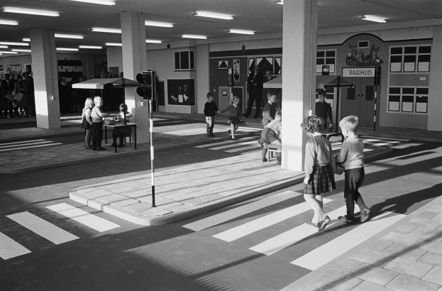Schwarz-weißes Foto von Kindern, die eine Straße an einer Ampel überqueren, mit Fußgängern in der Nähe, einem Verkehrssignalmast im Vordergrund und strukturellen Säulen mit Deckenleuchten im Hintergrund.