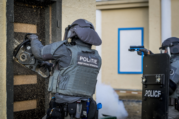 Zwei Polizisten in Schutzausrüstung stehen vor einem Gebäude, einer hält eine Waffe und der andere ein Objekt, mit einer Tür links und einer Tafel an der Wand im Hintergrund.