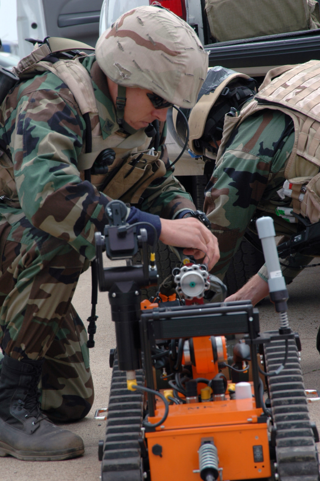 Zwei Männer in Tarnuniformen und Helmen arbeiten an einem kleinen Roboter auf dem Boden in einer militärischen Umgebung mit Fahrzeugen und Taschen im Hintergrund.