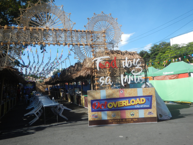 Eine KarnevalsSzene mit einem großen Riesenrad in der Mitte, umgeben von Hütten, Tischen, Stühlen, einer Schautafel, Dekorationen, Bäumen, Hochspannungsleitungen und einem bewölkten Himmel.