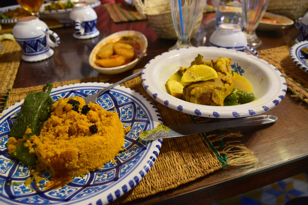 Ein Tisch mit Tellern, Gläsern, Schüsseln, Krügen, Besteck und Matten, mit im Hintergrund sitzenden Menschen bei einem traditionellen marokkanischen Essen.
