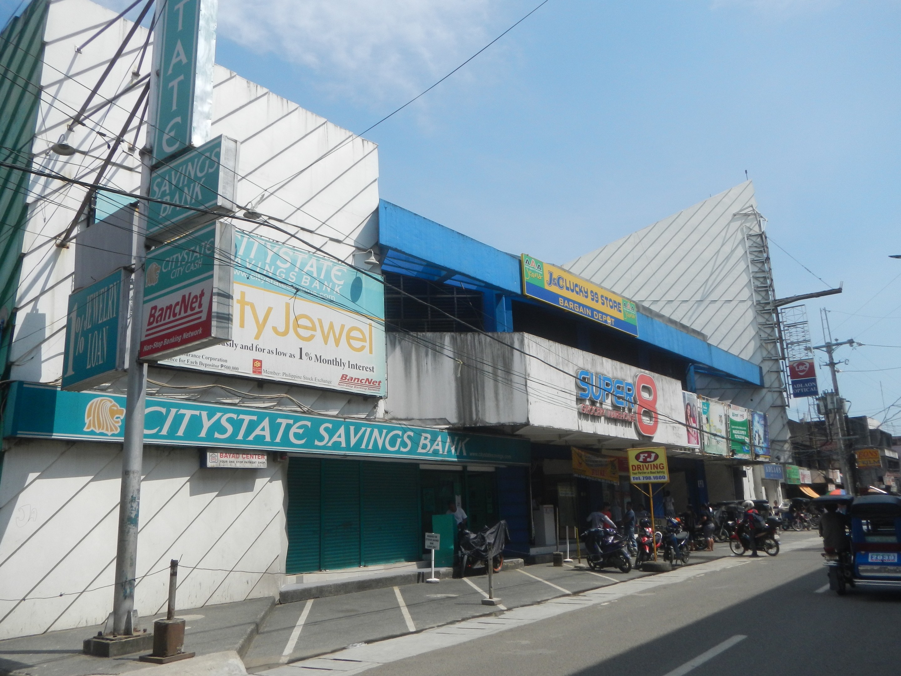 Eine belebte Stadtstraße mit Fahrzeugen, Fußgängern, Oberleitungen und Gebäuden, wobei die Citystate Savings Bank in Cebu City als Fokuspunkt unter einem bewölkten Himmel zu sehen ist.