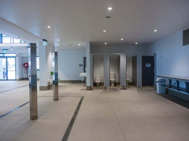 Große Öffentliche Toilette mit mehreren Toiletten, tragenden Säulen, einem Mülleimer, einer Tür, einem Schild, einem angebrachten Feuerlöscher und Deckenleuchten.