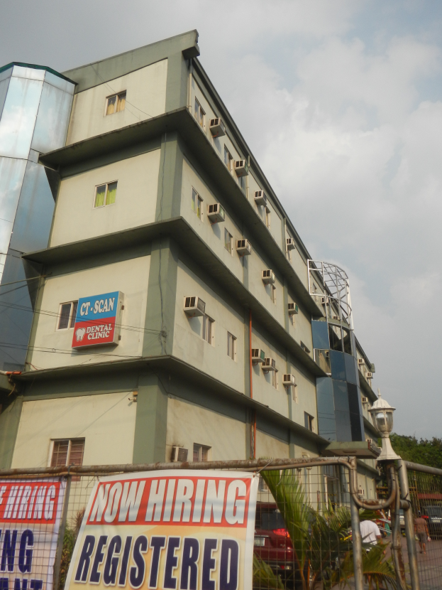 Ein Gebäude mit Fenstern, Klimaanlagen und einem "Jetzt Einstellen Registriert" Schild, umgeben von einem Metallzaun, einer Straßenlaterne, Pflanzen, Bäumen, Fahrzeugen, unter einem bewölkten Himmel.