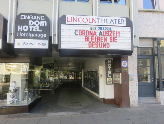 Das Lincoln Theater in Berlin, Deutschland, ist ein Gebäude mit Glasfenstern und -türen und einer Tafel mit Text darauf. Im Inneren gibt es verschiedene Objekte, die den Eindruck einer pulsierenden Stadtlandschaft vermitteln.