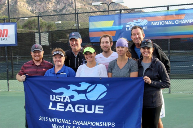 Eine Gruppe von Menschen auf einem Tennisplatz mit einem "2015 Usta National Championships"-Schild, einem Zaun, Bannern, Laternenpfählen, Bäumen, Bergen und einem klaren blauen Himmel im Hintergrund.