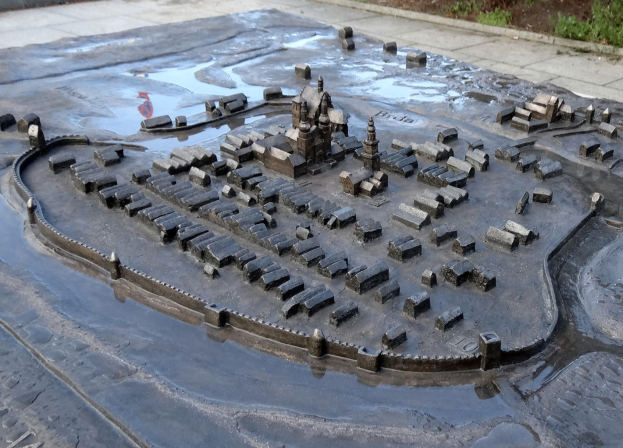 Eine Miniaturstadtmodell in einer Pfütze umgeben von Gebäuden, Grünflächen und einer Hintergrundwand.