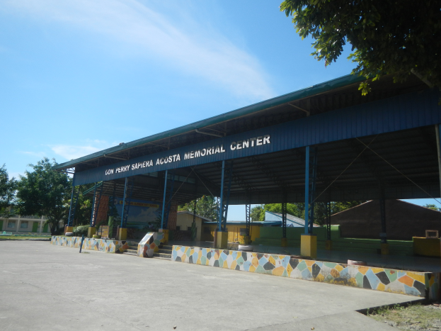Der Eingang zu einem Gebäude mit einer Tafel, auf der "Gedenkstätte" steht, umgeben von Bäumen, Häusern und einer Straße, mit einem Himmel voller weißer, flauschiger Wolken im Hintergrund.