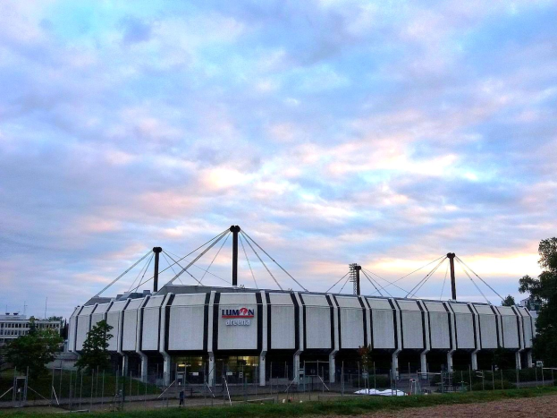 Großes Stadion mit grasigem Feld, Bäumen, Pfosten, Gebäuden und bewölktem Himmel im Hintergrund.