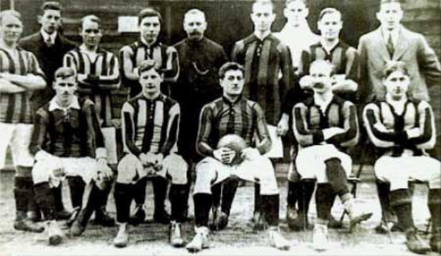 Schwarzes und weißes Foto einer Fussballmannschaft, einige stehen und einige sitzen, vor einer Wand mit dem Text «Ballymena Football Club» unten.