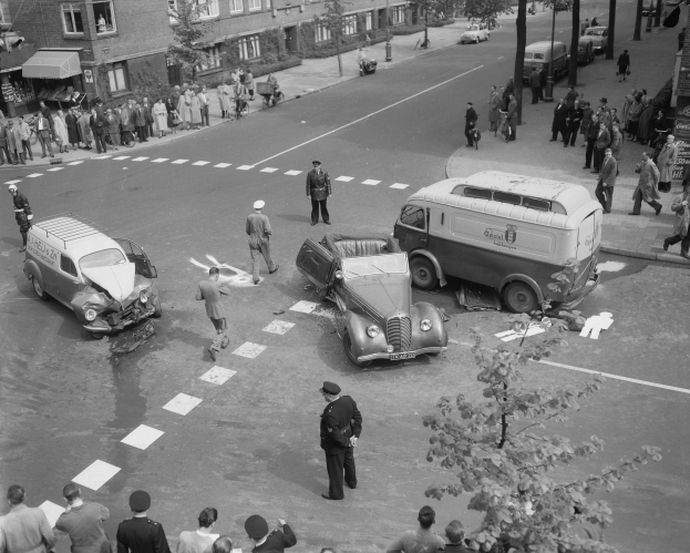 Eine Schwarz-Weiß-Szene eines Autounfalls an einer Straßenkreuzung, mit mehreren Fahrzeugen einschließlich eines Vans, einer Gruppe von Menschen drumherum und Gebäuden, Bäumen, Polen und Texttafeln im Hintergrund.