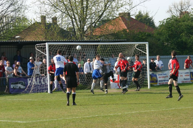 Fußballspieler spielen auf einem Feld mit einem Tor, während Zuschauer dahinter stehen, mit Bäumen und Häusern im Hintergrund.