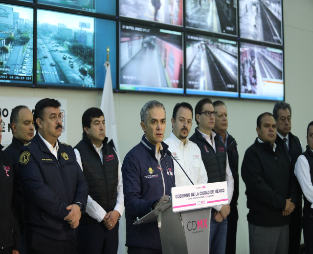 Gruppe von Männern, die vor einem Rednerpult mit einem Mikrofon und einer Tafel mit der Aufschrift 'Mexikanische Zoll- und Grenzschutzbehörde' stehen, mit Fernsehgeräten und einer Fahne an der Wand dahinter.