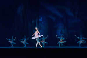 Eine Ballerina in rosa Tutu und Spitzenschuhen tanzt anmutig auf der Bühne, umgeben von anderen ähnlich gekleideten Tänzern, vor einem dunklen Hintergrund, der eine dramatische Atmosphäre schafft.