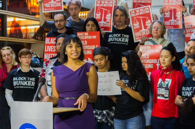 Eine Frau steht an einem Podium mit einem Mikrofon und einer Tafel und spricht zu einer Menge mit Schildern, mit Lichtern und Gegenständen im Hintergrund.