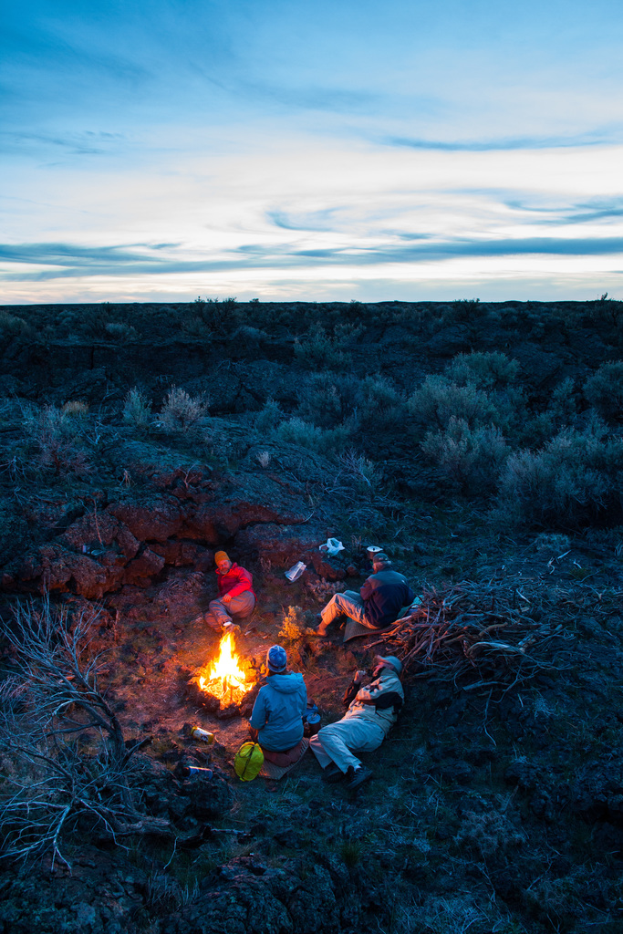 Ein Lagerfeuer mit Menschen, die darauf sitzen und liegen, mit Mützen tragend, mit einer Tasche und anderen Gegenständen auf dem Boden, umgeben von Bäumen und Himmel.