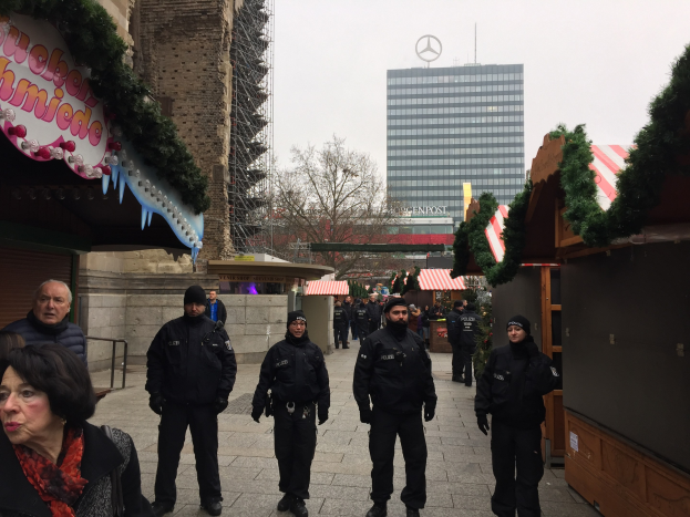Eine Gruppe von Polizisten in schwarzen Uniformen und Mützen steht vor einem geschmückten Weihnachtsmarkt in Berlin, mit festlichen Dekorationen, Gebäuden, Bäumen und einem klaren blauen Himmel im Hintergrund.
