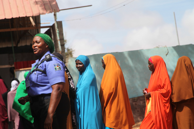 Gruppe von Frauen, die vor einem Gebäude stehen, mit einer Polizistin im Vordergrund, Schuppen, Pfähle, Drähte, Bäume und einen klaren blauen Himmel im Hintergrund.
