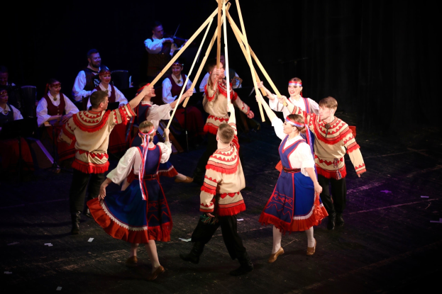 Bunt kostümierte Tänzer weben Bänder um einen Maibaum, während Musiker im Hintergrund spielen.