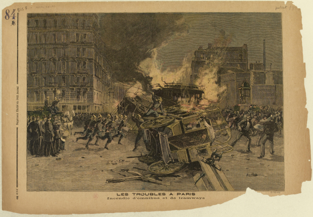 Eine Gruppe von Menschen steht um ein brennendes Auto in der Mitte einer Straße, mit Gebäuden im Hintergrund und Text am unteren Bildrand.