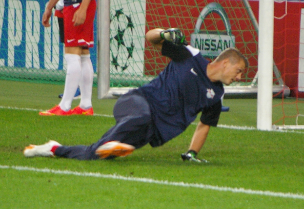 Ein Fussballspieler in Handschuhen taucht ab, um einen Ball vor einem Tor zu fangen, mit zwei Personen, einem Netz und einer Banner mit Text im Hintergrund.