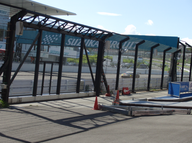 Eine Rennstrecke mit geparkten Autos, Verkehrskegeln, einem Zaun, einem Schuppen, einer Fahne, Pflanzen, Bäumen und einem bewölktem Himmel.