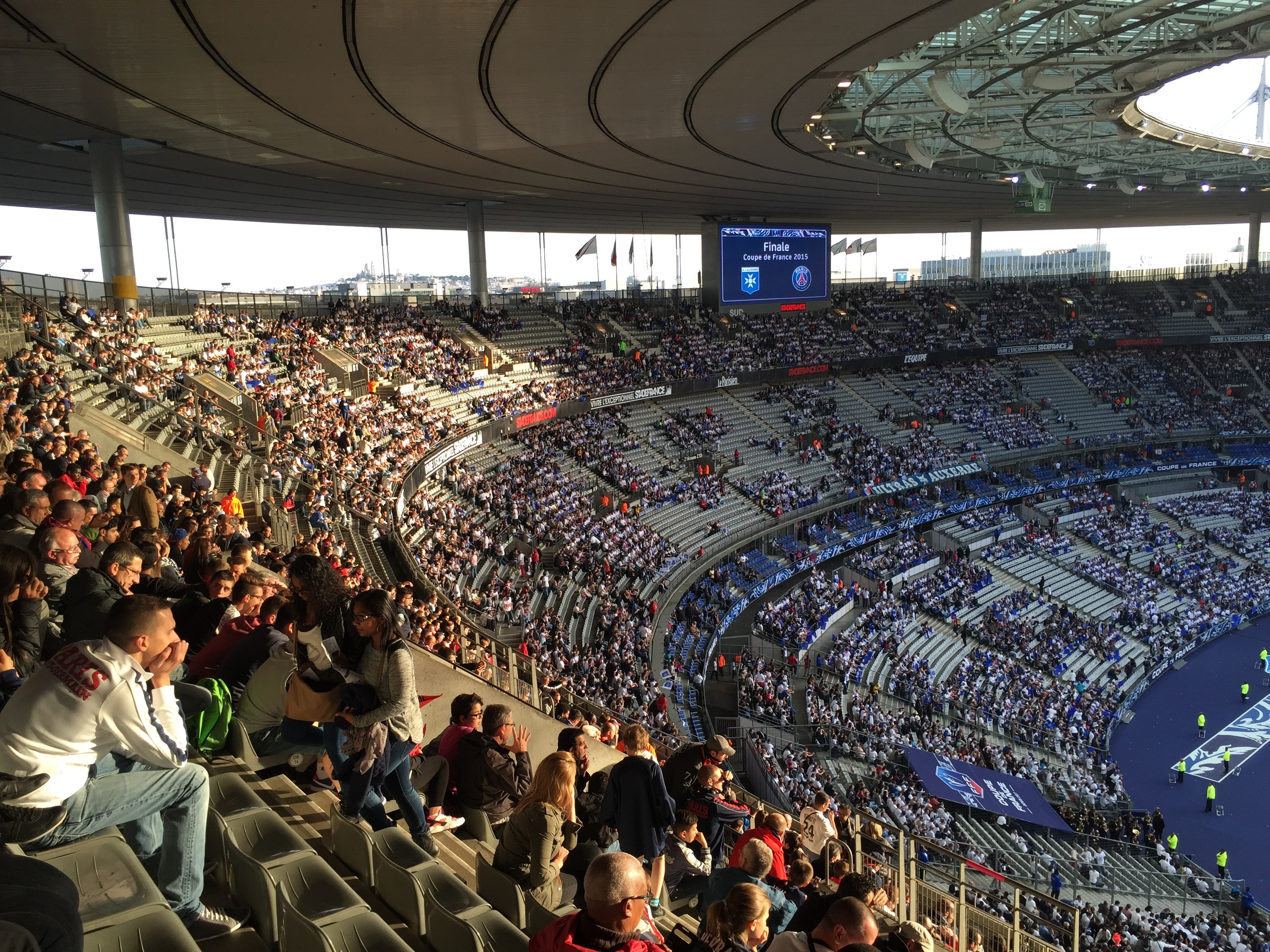 Große Menschenmenge in einem Stadion bei einem Fußballspiel mit einer Bühne rechts, Fahnen, Stangen, einem Bildschirm und dem Allianz Arena in München, Deutschland im Hintergrund.