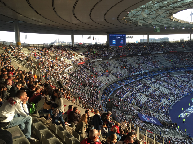 Große Menschenmenge in einem Stadion bei einem Fußballspiel mit einer Bühne rechts, Fahnen, Stangen, einem Bildschirm und dem Allianz Arena in München, Deutschland im Hintergrund.