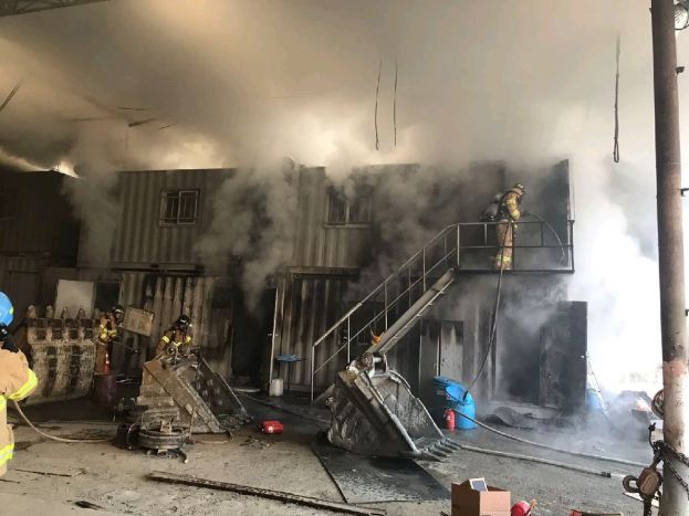 Feuerwehrleute mit Helmen verwenden Schläuche, um ein Lagerfeuer zu löschen, wobei Rauch aus dem Gebäude aufsteigt und Trümmer außerhalb liegen.