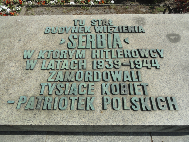 Denkmalstein mit einer Plakette vor einem Gebäude umgeben von blühenden Pflanzen.