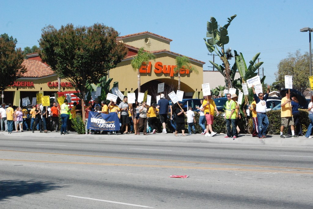 Eine Gruppe von Menschen, die mit Schildern und einer Fahne durch die Natur gehen, mit kleinen Büschen, einem Gebäude mit Namensschildern und Fahrzeugen im Hintergrund.
