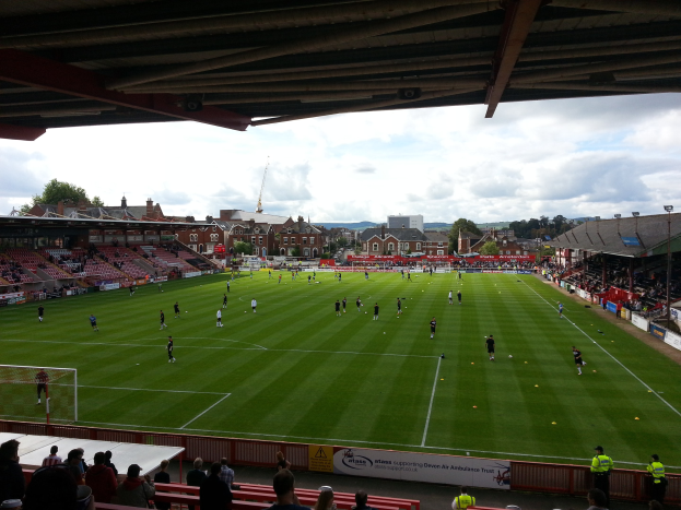 Ein Fußballspiel im St. Mary's Stadium, mit Zuschauern auf den Tribünen und Stehplätzen, einem Torpfosten auf der linken Seite und im Hintergrund Gebäude, Bäume und eine bewölkte Himmel.