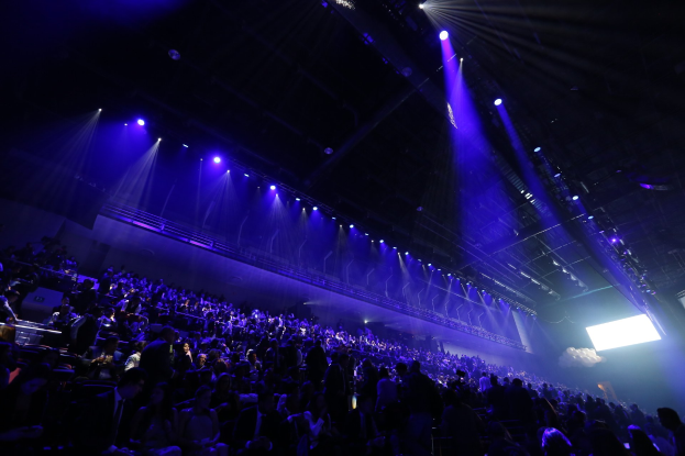 Eine große Menschenmenge sitzt einer Bühne mit Deckenbeleuchtung gegenüber, und auf der rechten Seite ist ein Bildschirm zu sehen.