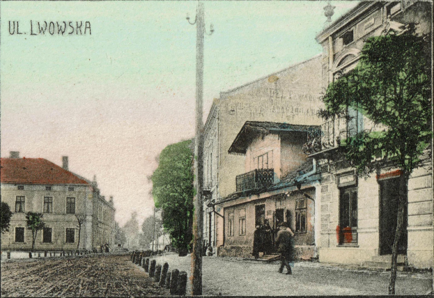 Ein altes Foto einer Straße in Uluwowska, Polen, mit Gebäuden, Bäumen, Pfählen, Menschen, einer Straße im Vordergrund und dem Himmel im Hintergrund, mit Text oben auf dem Bild.