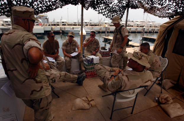 Gruppe von Männern in Mützen und Schwimmbrillen sitzt um einen Tisch auf einem Boot, mit Taschen auf dem Boden und einem Zelt rechts; Boote, Stangen, Gebäude und ein klarer blauer Himmel im Hintergrund.