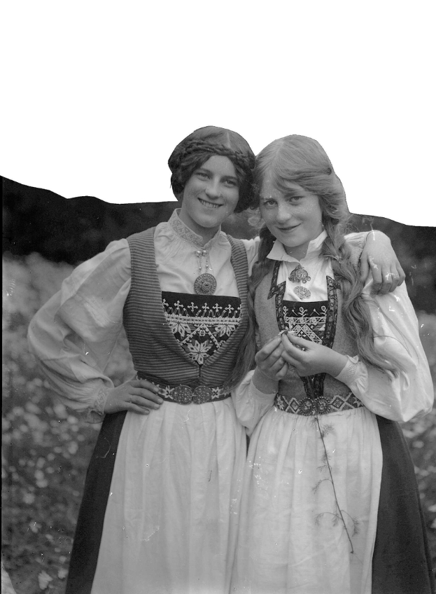 Zwei junge Frauen in traditioneller bayrischer Tracht, mit weißen und schwarzen Kleidern mit aufwändiger Stickerei und Perlen, posieren vor der Kamera gegen einen Hintergrund aus üppigen grünen Pflanzen und einem hellen weißen Himmel.