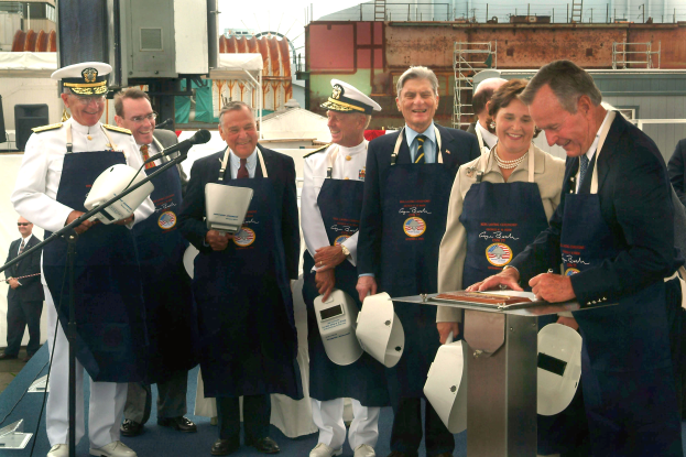Eine Gruppe von Menschen vor einem Rednerpult, die lächeln und Helme halten, mit einem Mikrofonständer links und einem Gebäude im Hintergrund.